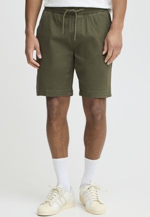 Olijfgroene shorts met een elastische tailleband en trekkoorden. Rechte snit, reguliere lengte, gemaakt van katoenen stof. Draagt witte sokken en schoenen.