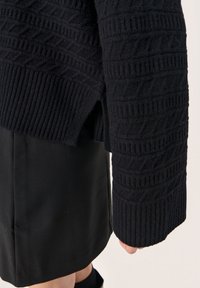 Czarny sweter z dzianiny o fakturowanym wzorze, z prążkowanymi mankietami i bocznymi rozcięciami, zestawiony z dopasowaną czarną spódnicą. Miękki, ciepły materiał.