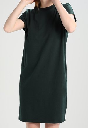 Femme portant une robe T-shirt unie de couleur vert foncé, à manches courtes et longueur au genou, sur un fond neutre.