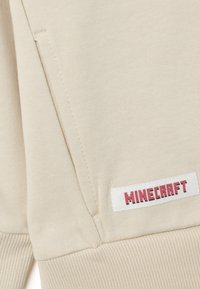 Felpa beige con texture a costine, vestibilità aderente e un'etichetta bianca con il testo rosso "MINECRAFT" sul lato.