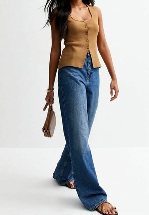 Femme marchant portant un haut marron sans manches, un jean bleu à jambes larges, des sandales et portant un petit sac à main marron.