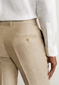 Beżowe lniane spodnie z tylną kieszenią zapinaną na guzik i teksturowanym wykończeniem, w zestawie z białą koszulą z długim rękawem.