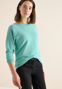 Turkusowy sweter z prążkowanego materiału z dekoltem w łódkę i rękawami 3/4, w zestawie z czarnymi spodniami; ma luźny krój i teksturowany materiał.