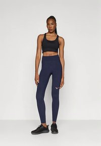 Nő áll navy színű leggingsben, rózsaszín logóval, fekete sportmelltartóban és fekete sportcipőben, egyszínű világos háttér előtt.