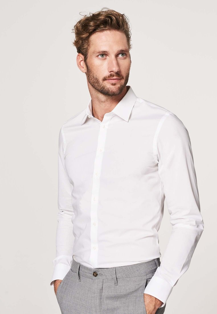 Camicia bianca a maniche lunghe con colletto classico e chiusura a bottoni. Tessuto morbido; design aderente. Abbinata a pantaloni grigi chiari.