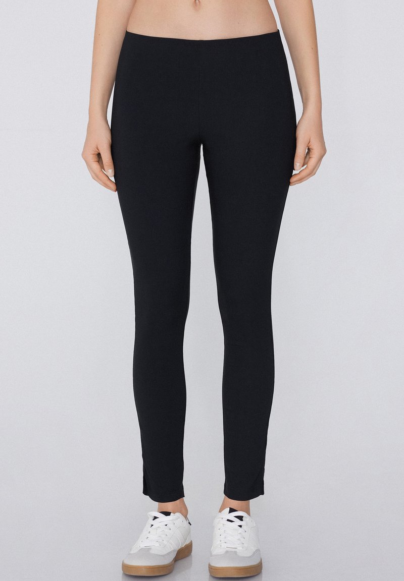 Person trägt schwarze Slim-Fit-Hose und weiße Sneakers, steht vor hellem Hintergrund und zeigt nur den Unterkörper.