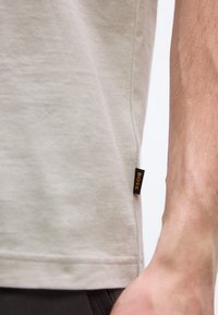 Κοντινό πλάνο της πλαϊνής ραφής σε μπεζ T-shirt με μαύρη ετικέτα που γράφει "BOSS" δίπλα στο γυμνό χέρι ενός ατόμου.