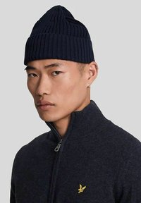 Berretto a coste blu navy con cima appuntita. Indossato da un modello in un maglione zip-up scuro con un piccolo logo giallo sul petto.