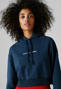 Žena s kučeravými vlasmi, oblečená v tmavomodrej skrátenej mikine s nápisom "Red Bull Racing" a červených nohaviciach, čelom vpred na jednoduchom pozadí.