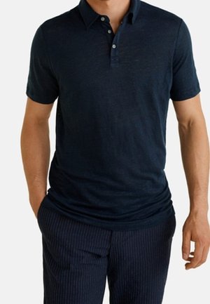Mann trägt ein dunkelblaues kurzärmeliges Poloshirt und dunkelblaue Nadelstreifenhose, Hände entspannt an den Seiten.