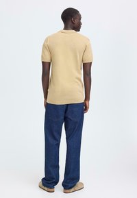 Polo en maille beige à manches courtes porté avec un pantalon en jean denim foncé ample et des chaussures marron clair sans lacets. Couture visible sur les bords du polo.