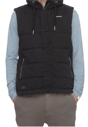 Mann trägt schwarze, gesteppte ärmellose Kapuzenweste über hellblauem Langarmhemd mit den Händen in grauen Hosentaschen.