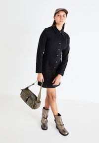 Μαύρο τζιν φόρεμα με μακριά μανίκια και κουμπιά μπροστά, συνδυασμένο με μεταλλικά καφέ ελαστικά μποτάκια και μια ελαιοπράσινη τσάντα χιαστί.