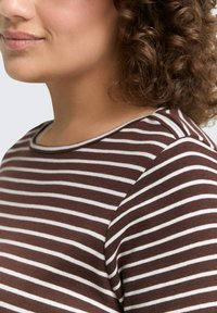 Frau mit lockigem Haar, die ein braun-weiß gestreiftes Hemd trägt, zu sehen von Hals bis zur oberen Brust und Schulter.