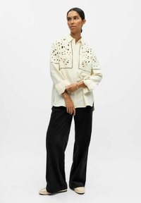 Helle beige Bluse mit Auschnitten in floralen Mustern an den Schultern, kombiniert mit schwarzen Cordhosen im weiten Schnitt und beigen Slippern.
