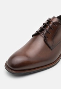 Chaussure habillée en cuir marron avec une finition polie, un bout arrondi et un système de laçage à cinq œillets. Présente des coutures discrètes et une semelle contrastée.