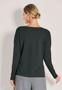 Frau mit blonden Haaren in einem tiefen Dutt, trägt große goldene Ohrringe, einen dunkelgrünen Langarmpullover und graue Hosen und schaut von der Kamera weg.