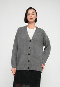 Γκρι ριγωτή ζακέτα με V-λαιμόκοψη, πέντε μαύρα κουμπιά στο μπροστινό μέρος και μακριά μανίκια, σε συνδυασμό με λευκό τοπ και μαύρη φούστα.