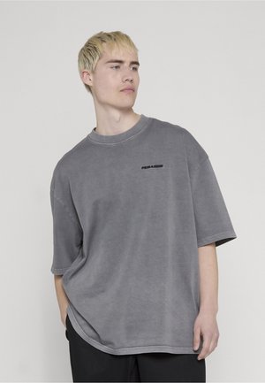 Młody mężczyzna z krótkimi blond włosami, ubrany w luźną szarą koszulkę z małym czarnym napisem na piersi, stojący na tle jednolitego jasnego tła.