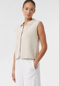 Beżowa bezrękawnik dzianinowy z kołnierzykiem, z przodu zapinany na guziki i o pudełkowym kroju, w połączeniu z białymi luźnymi spodniami. Teksturowany materiał.