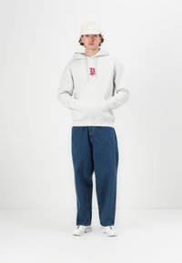 Felpa grigia con tasca frontale e patch del logo, abbinata a jeans blu larghi e scarpe da ginnastica bianche. Il modello indossa un cappellino chiaro.
