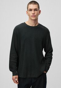 Czarna koszula z długim rękawem o teksturowanym, prążkowanym wzorze, z okrągłym dekoltem i luźnym krojem. Noszona z ciemnymi dżinsami.