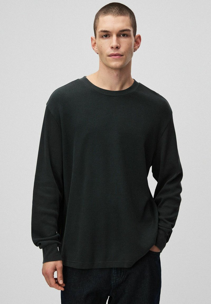 Czarna koszula z długim rękawem o teksturowanym, prążkowanym wzorze, z okrągłym dekoltem i luźnym krojem. Noszona z ciemnymi dżinsami.