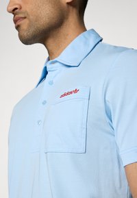 Light blue polo shirt with a collar, featuring a red embroidered logo on the chest pocket. Made of soft, lightweight fabric.