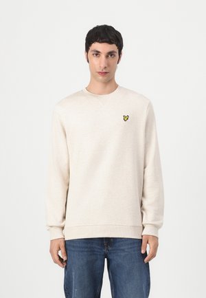 Homme debout portant un sweat-shirt crème à col rond avec un petit logo d'oiseau jaune sur la poitrine et un jean bleu, sur un fond uni.
