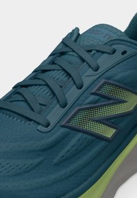 Μπλε αθλητικό παπούτσι με σκούρα μπλε κορδόνια και πράσινο λογότυπο "N" στο πλάι, με υφασμάτινο επάνω μέρος με υφή και μαλακή σόλα.