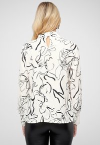 Blouse à manches longues en tissu crème avec un motif floral abstrait noir, dotée d'un détail dos en œillet, d'une fermeture à bouton et de poignets ajustés.