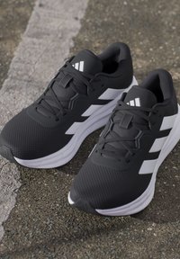 Chaussures de course Adidas noires et blanches avec lacets, placées sur un bitume texturé près d'une ligne blanche fanée.