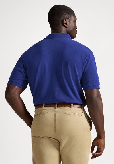 Mann som står med ryggen til, iført en blå kortermet poloshirt, beige bukser og et brunt belte mot en ensfarget hvit bakgrunn.