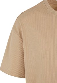 Chemise beige à manches courtes avec une texture côtelée et un col rond, montrée de l'épaule jusqu'au milieu du torse sur fond blanc.