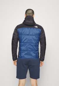 Puffig jacka i mörkblått och svart, med huva, som har en slät yta och elastiska muddar. Logotyp på bakre axel.