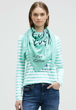 Young woman wearing a mint green striped long-sleeve shirt and matching scarf with "Limited Edition" text, hands in dark blue jeans pockets.