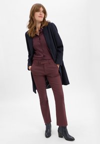 Bordeauxkleurige blouse met knopen, bijpassende broek met riemlussen, in laagjes onder een navy wollen jas. Zwarte enkellaarsjes met hak maken de outfit compleet.