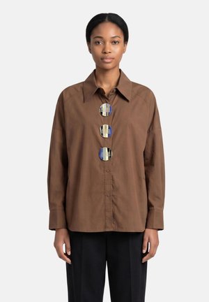 Femme portant une chemise marron à manches longues avec de gros boutons ronds décoratifs sur le devant et un pantalon noir sur un fond uni.