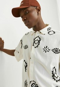 Košulja kratkih rukava, s dugmadi, u off-white tkanini, s crnim cvjetnim uzorkom. Model nosi smeđu kapu, s rukom naslonjenom na zid.