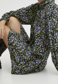 Svart blommig klänning med lila blommor och gröna blad. Har långa ärmar, krage och en lös passform, tillverkad av lätt tyg.