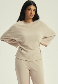 Jasno beżowy, oversize sweter i dopasowane spodnie wykonane z miękkiego materiału, z falbankami na rękawach i u dołu. Prosty, luźny fason.