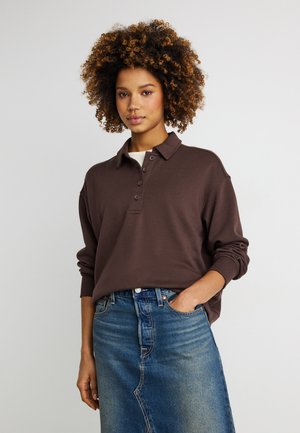 Young woman with curly hair wearing a brown long-sleeve polo shirt and a blue denim skirt, standing with one hand in her pocket.