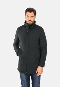 Cappotto impermeabile nero con colletto alto, chiusura a bottoni automatici e tasche laterali. Realizzato in un tessuto liscio con un design aerodinamico.