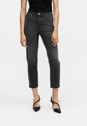 Kvinde iført sorte, cropped jeans med lige ben, sort ærmeløs top og sorte spidse høje hæle med ankelstropper.
