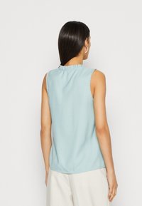 Top sin mangas de color azul claro con un escote con volantes. Textura suave, dobladillo redondeado y un diseño sencillo. La vista trasera muestra detalles minimalistas.