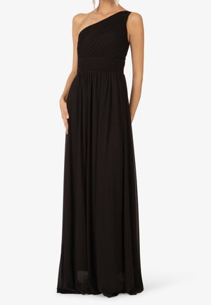 Kvinde iført en lang sort asymmetrisk aftenkjole med plisseret bodice og flydende nederdel, stående med den ene hånd i siden.