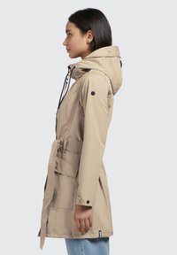 Beige rain jacket with a drawstring hood, belt, and side pockets. Features a smooth texture and button accents, styled with a relaxed fit.