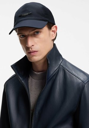 Giacca in pelle blu navy con collo alto, cerniera argento e maglione girocollo grigio sotto. Cappello blu navy scuro con dettaglio logo in metallo.