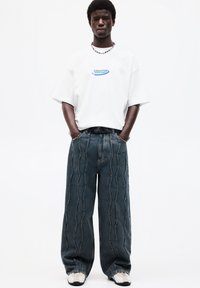 White oversized t-shirt with blue graphic text, paired with wide-leg dark denim jeans featuring stitched wave patterns and a multicolored belt.