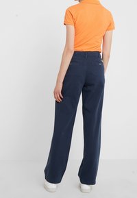 Orange pikétröja med korta ärmar, kombinerad med mörkblå vida byxor. Visar en mjuk textur och avslappnad passform. Vita sneakers kompletterar utseendet.